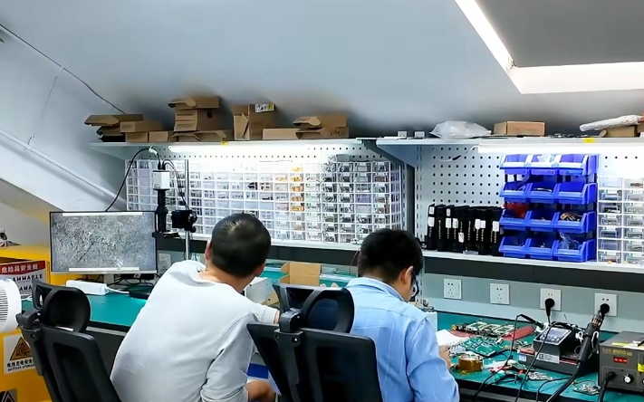 Personas trabajando en electrónica en una mesa con un almacén de componentes al fondo